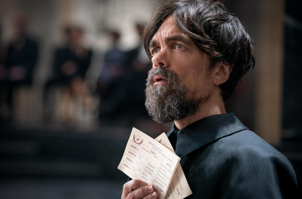 Peter Dinklage as Dean Casca Highbottom holding a mentor assignment card in The Hunger Games prequel.